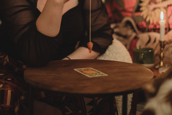 Vägledaren håller en pendel i högra handen som pendlar över ett tarotkort som ligger på ett bord. Mediala tjänster är för dig som önskar andekontakt.